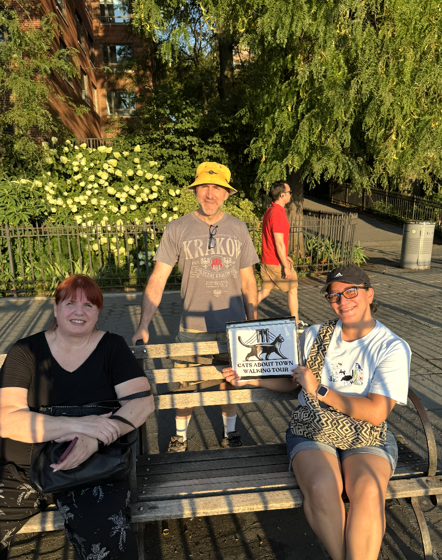 Three tour guests on a park bench holding the Cats About Town Walking Tour sign in evening light